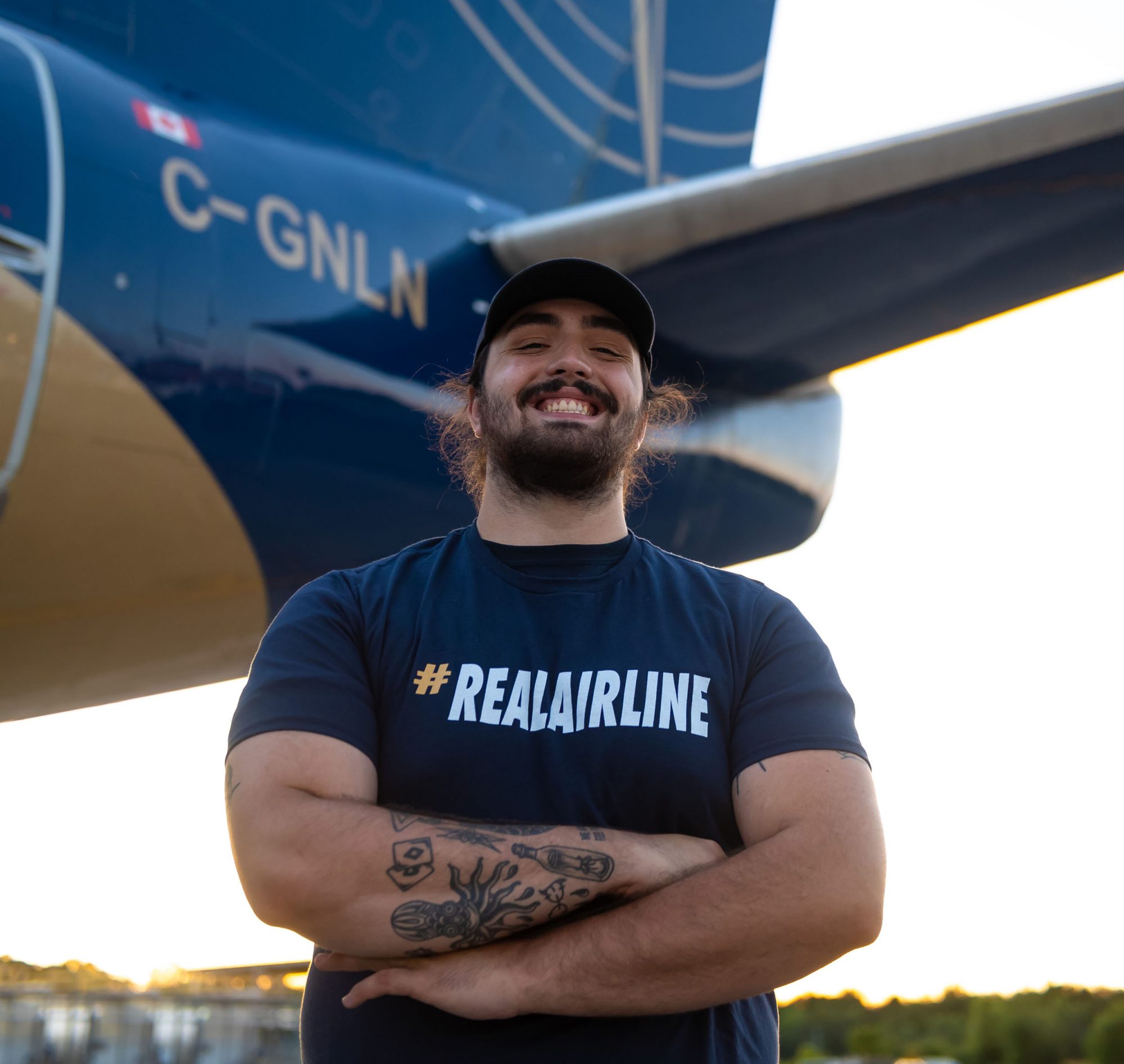 Homme souriant en t-shirt #REALAIRLINE et casquette, debout devant la queue d’un avion bleu avec l’immatriculation C-GNLN.
