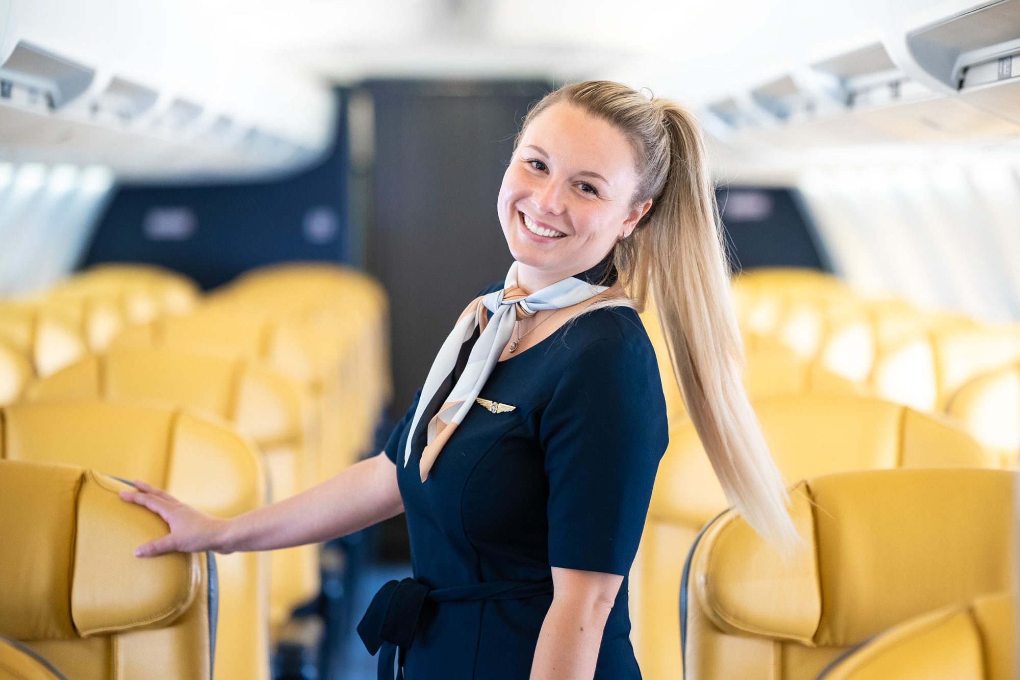 Agente de bord souriante en uniforme bleu marine dans une cabine d’avion aux sièges jaunes.