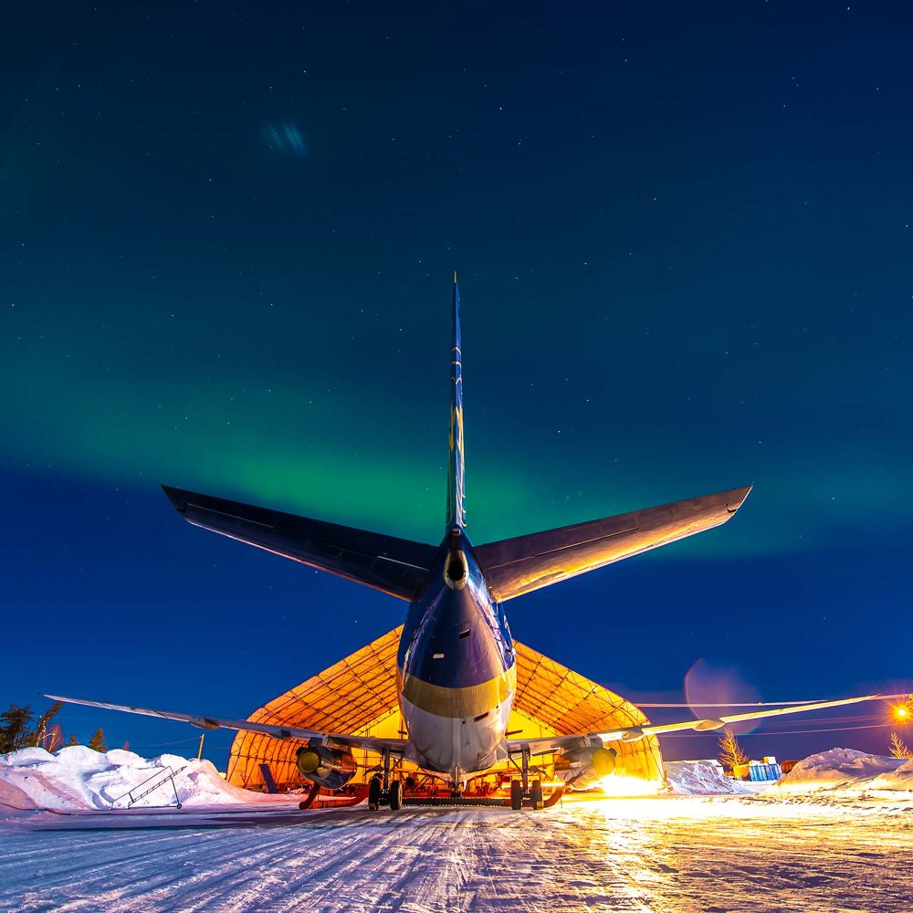 Avion vu de l’arrière devant un hangar éclairé, avec aurores boréales dans le ciel nocturne enneigé.